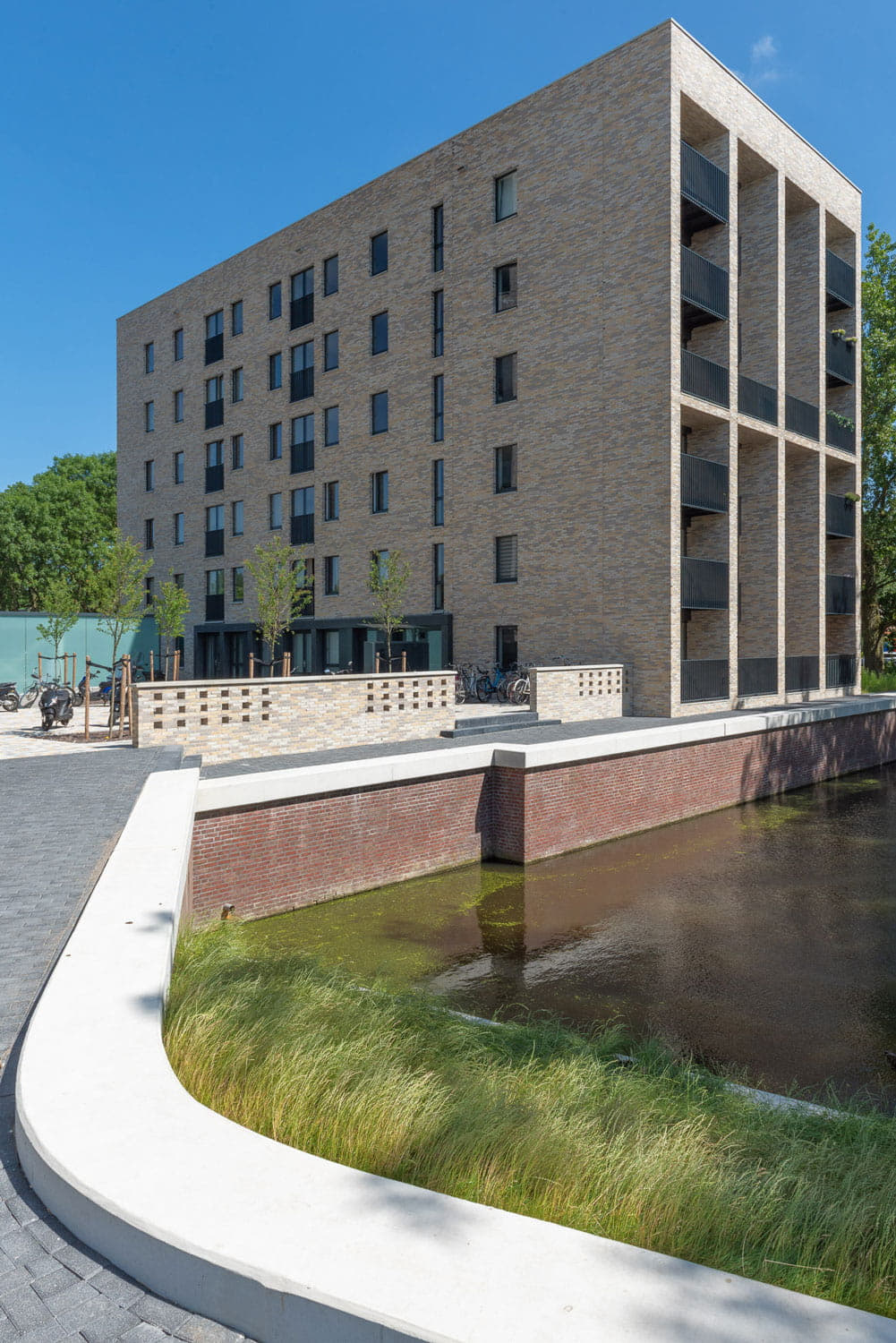 hoog gebouw met lichte bakstenen en steenstrips met op de voorgrond een brug